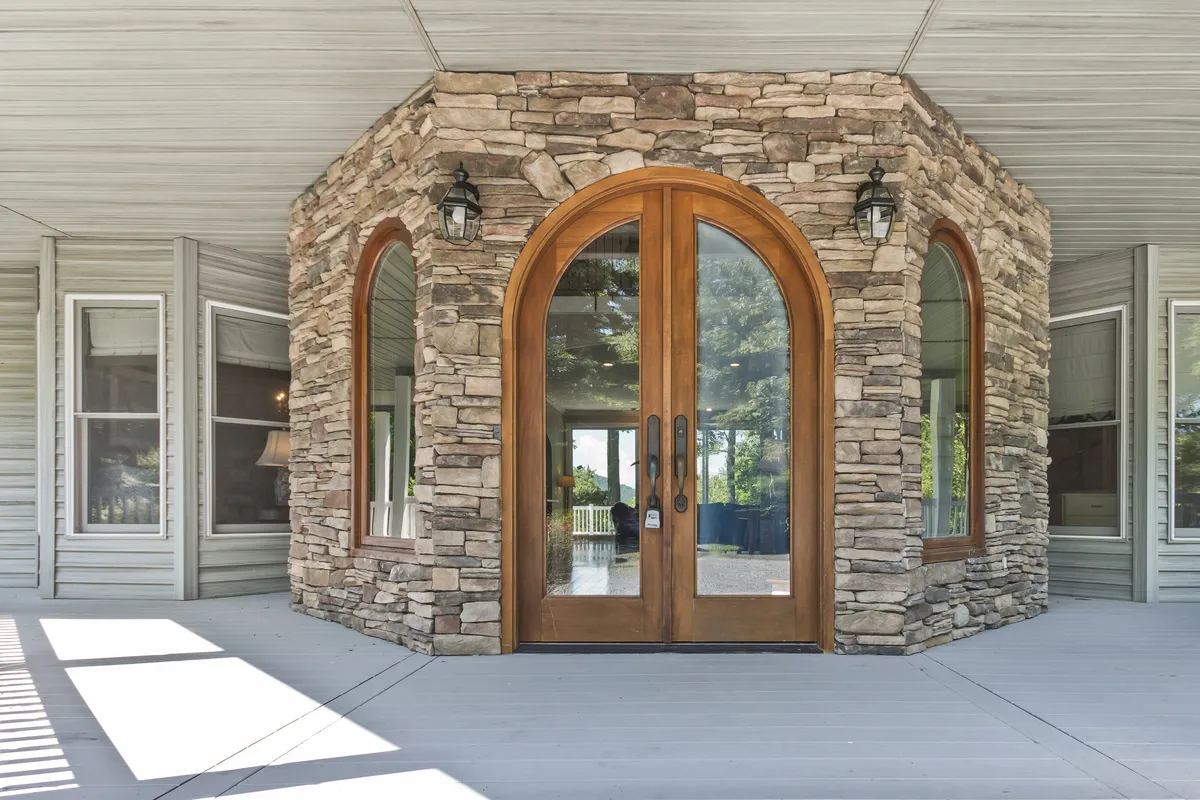 The image features the exterior of the property, showcasing impressive arched wooden doors, a classic stone facade, and expansive windows. The lush landscaping and spacious porch invite guests to imagine both arrival and relaxation, offering a sense of place and welcome.