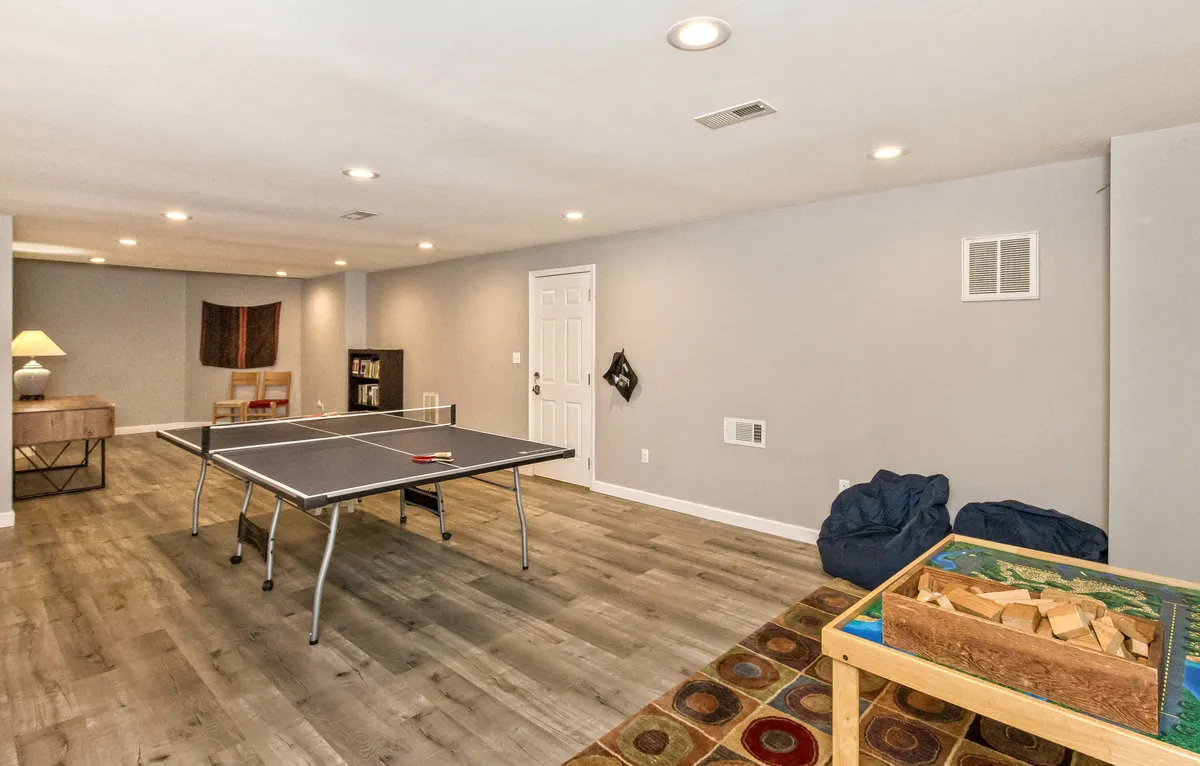 This image showcases a lively basement playroom featuring a ping pong table at the center, surrounded by colorful bean bag chairs and a bright area rug. The wooden storage table suggests space for games and creative activities, capturing the essence of family fun.