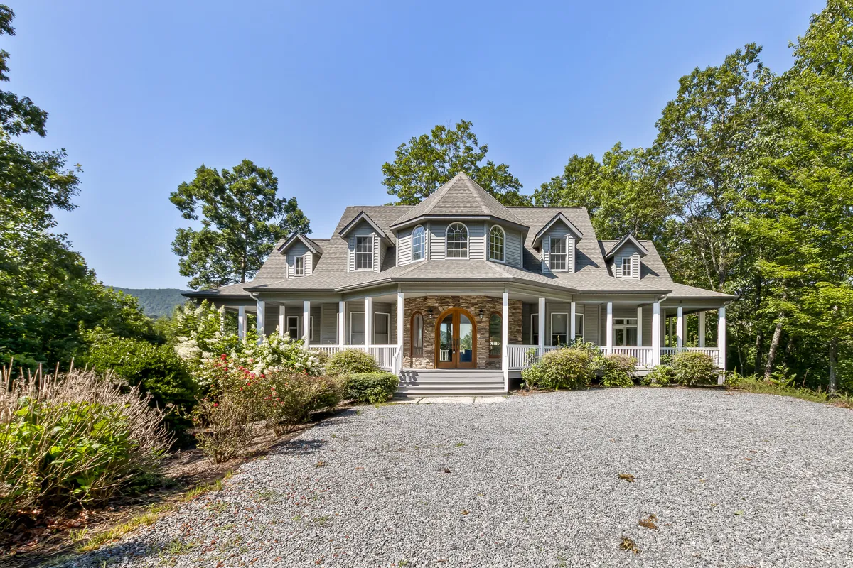 The image showcases the grand exterior of a spacious mountain home with a large, inviting front porch framed by arched double doors and dormer windows. The home is nestled among lush greenery, hinting at a serene natural setting and offering glimpses of sunset views and possible wildlife sightings. The porch is designed for relaxation and gathering, perfect for families or groups to enjoy the outdoors together.