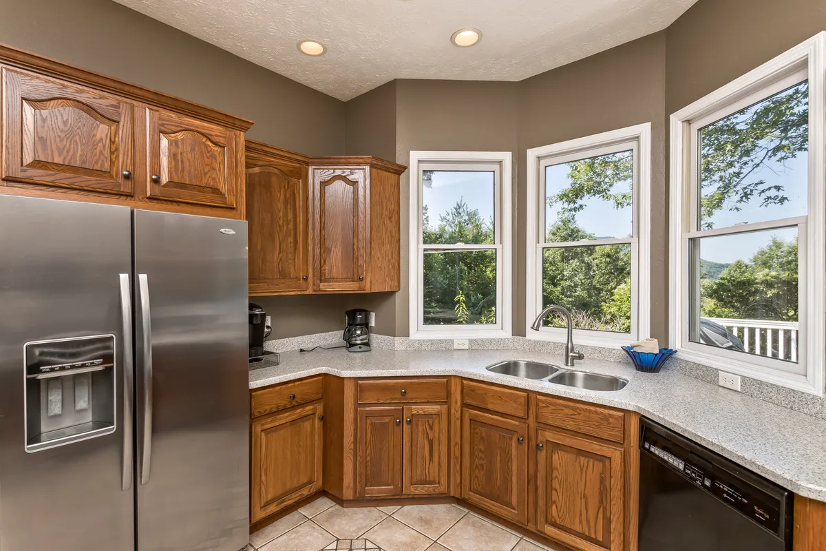 A spacious, sun-drenched kitchen with a granite countertop, stainless steel refrigerator, kitchen sink, and cabinets with a warm wood finish. Large windows offer scenic views and bring in natural light, creating an inviting atmosphere for meal prep and family gatherings. The countertop is uncluttered, highlighting the kitchen’s readiness for gourmet cooking or casual breakfasts.