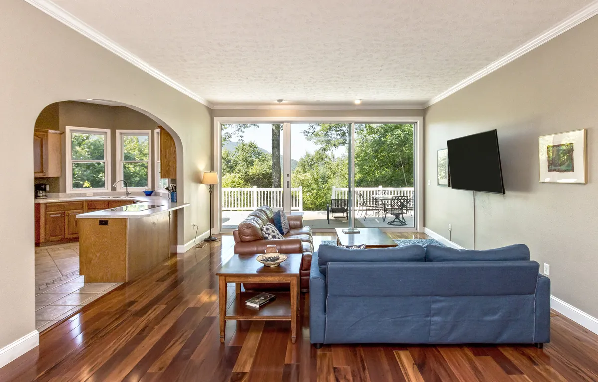 A stylish living area with expansive windows overlooking greenery, a modern TV, and a comfortable seating area. The open layout connects to a visible kitchen space through an archway, reinforcing the open-plan concept for easy movement and togetherness.