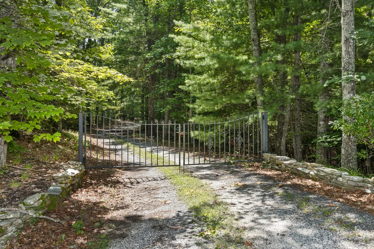 A tranquil gated entrance set amidst tall trees and a gravel path leading into the heart of nature. The setting suggests peaceful seclusion, easy access to walking trails, and the adventure of exploring the property and surrounding forests with pets.